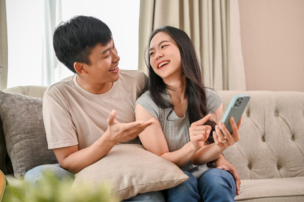 Happy young Asian couple, boyfriend and girlfriend, enjoy talking while using a smartphone together on a sofa in the living room.