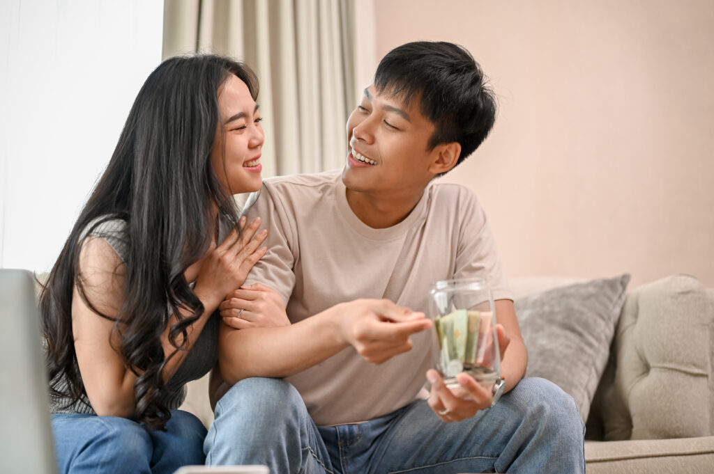 A lovely, happy young Asian new married couple is planning their savings together in the living room, putting coins and some money in a jar.