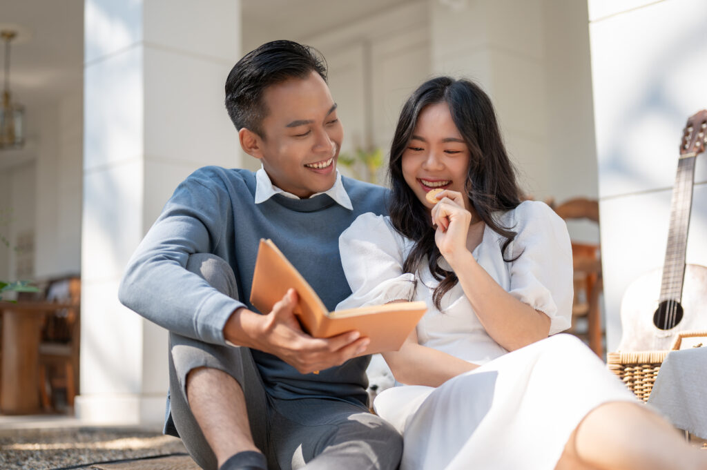 Lovely sweet young Asian couple enjoying picnic on a sunny day at home together, reading a book, having fun , enjoying talking.