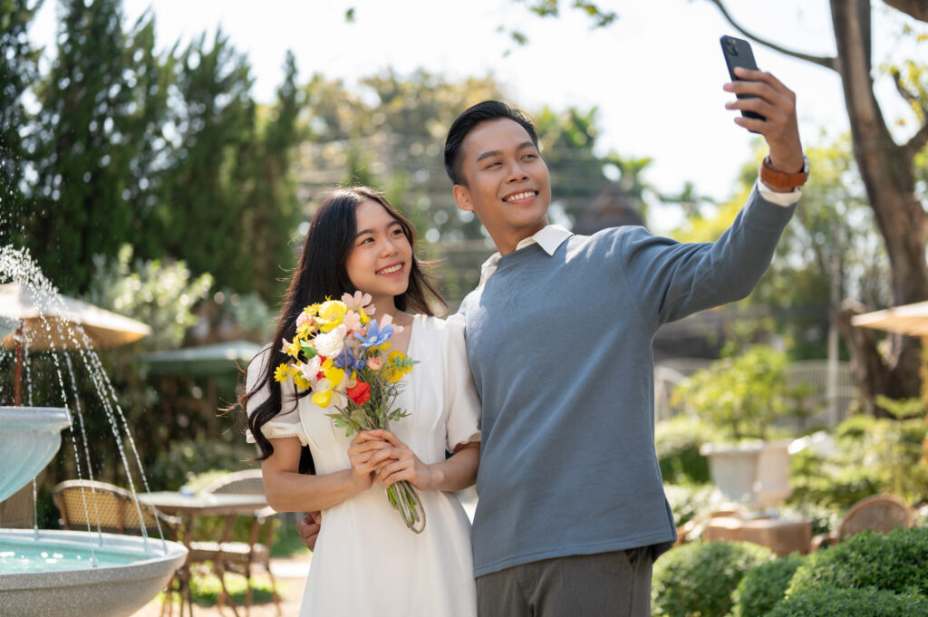 A cheerful young Asian man surprises his girlfriend with beautiful flowers, taking selfies together in a beautiful garden. romantic couple, bonding, anniversary, love